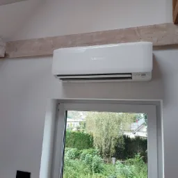 Biały klimatyzator Mitsubishi zamontowany na ścianie pod drewnianą belką stropową, widok z okna na zieleń.