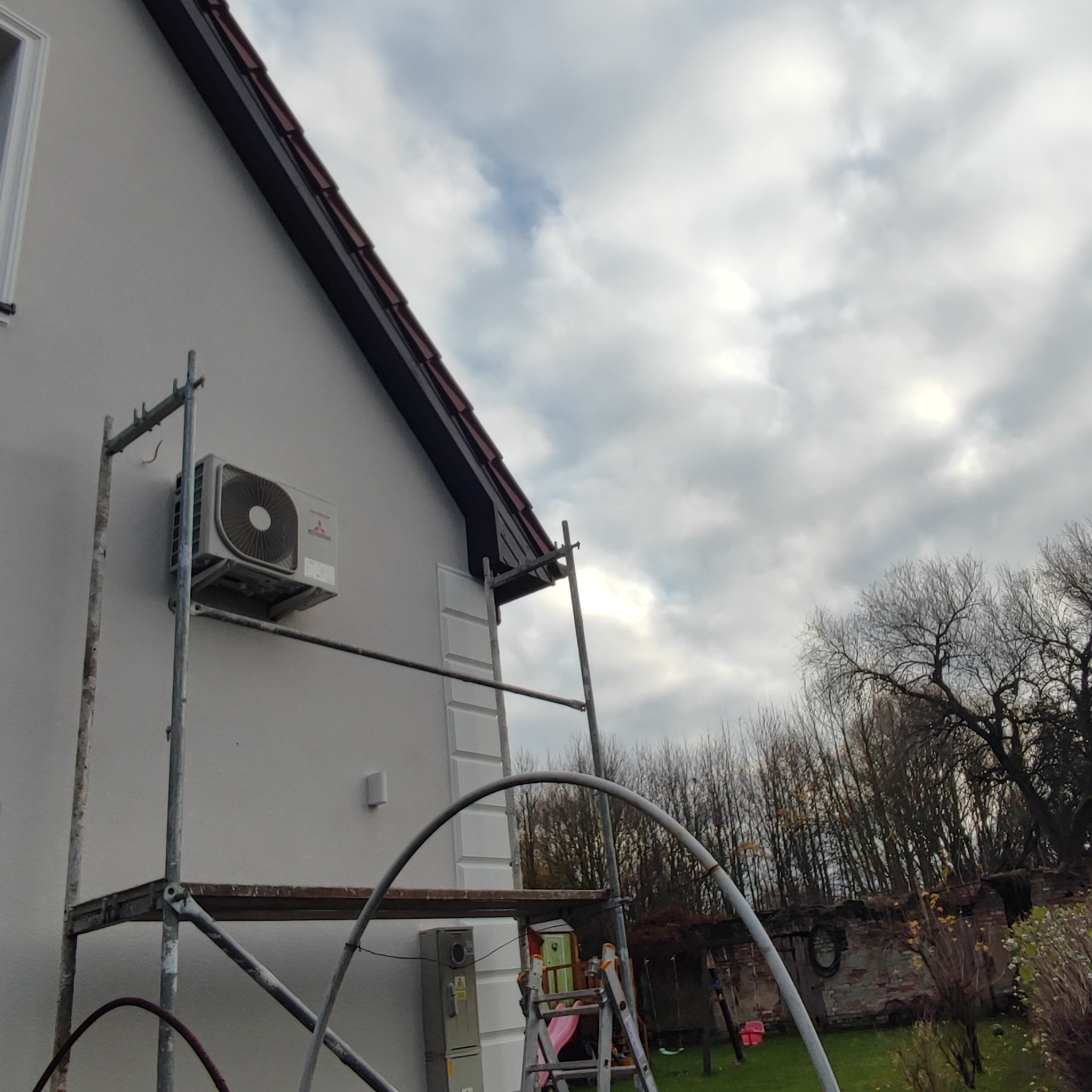 Montaż klimatyzacji zewnętrznej na elewacji budynku, widoczne rusztowanie i drabina, pochmurne niebo w tle.