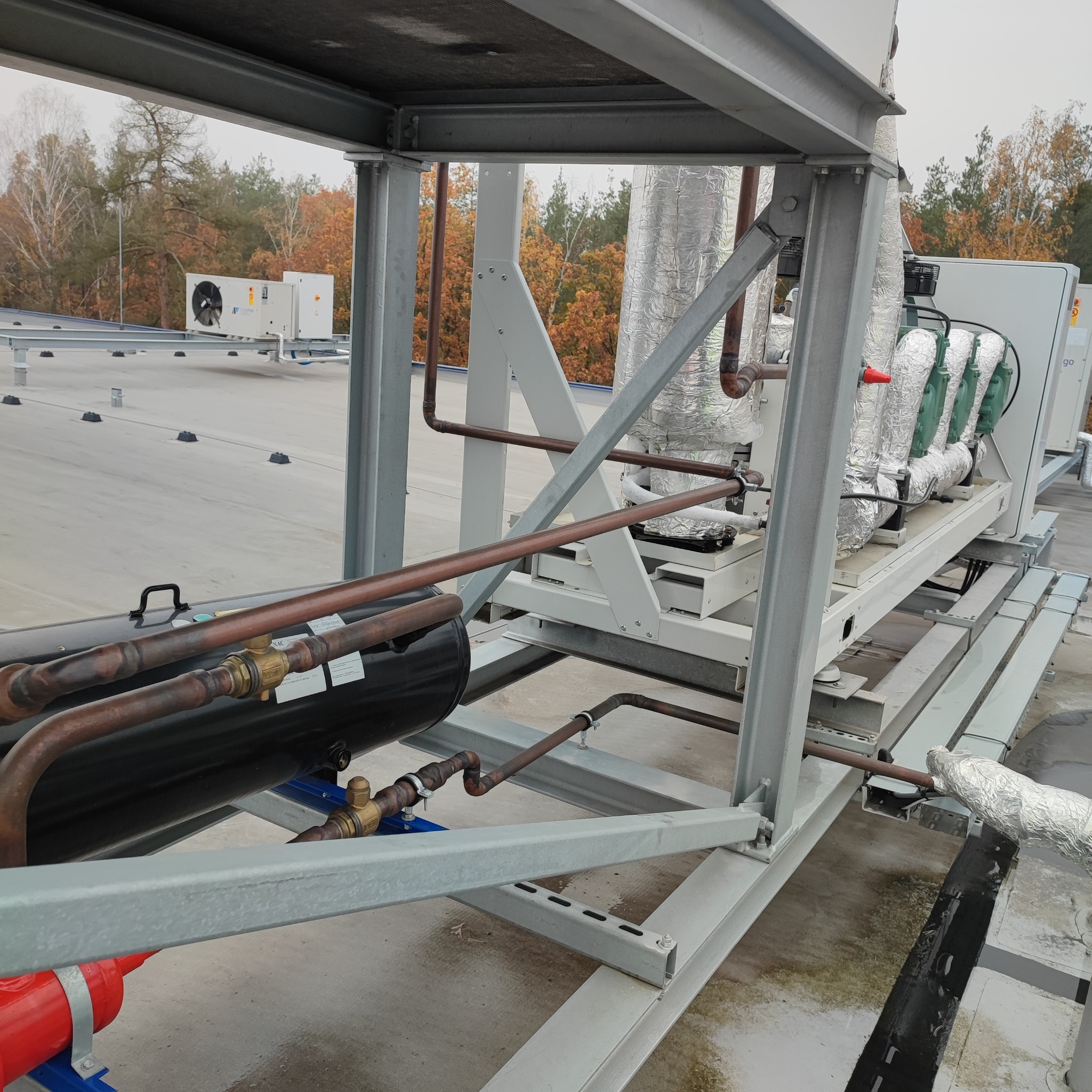 Instalacja chłodnicza na dachu budynku, widoczne rury miedziane, zbiornik ciśnieniowy, klimatyzatory i konstrukcja wsporcza z metalu.