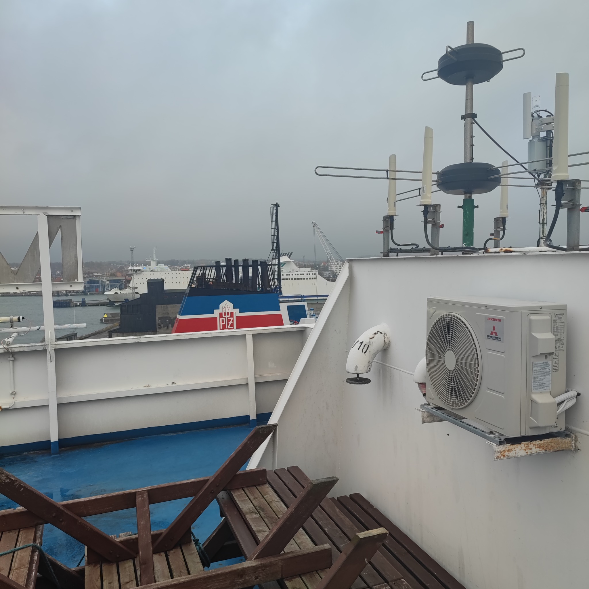 Klimatyzator Mitsubishi zamontowany na białej ścianie na statku, w tle widok na port i inne statki w pochmurny dzień.