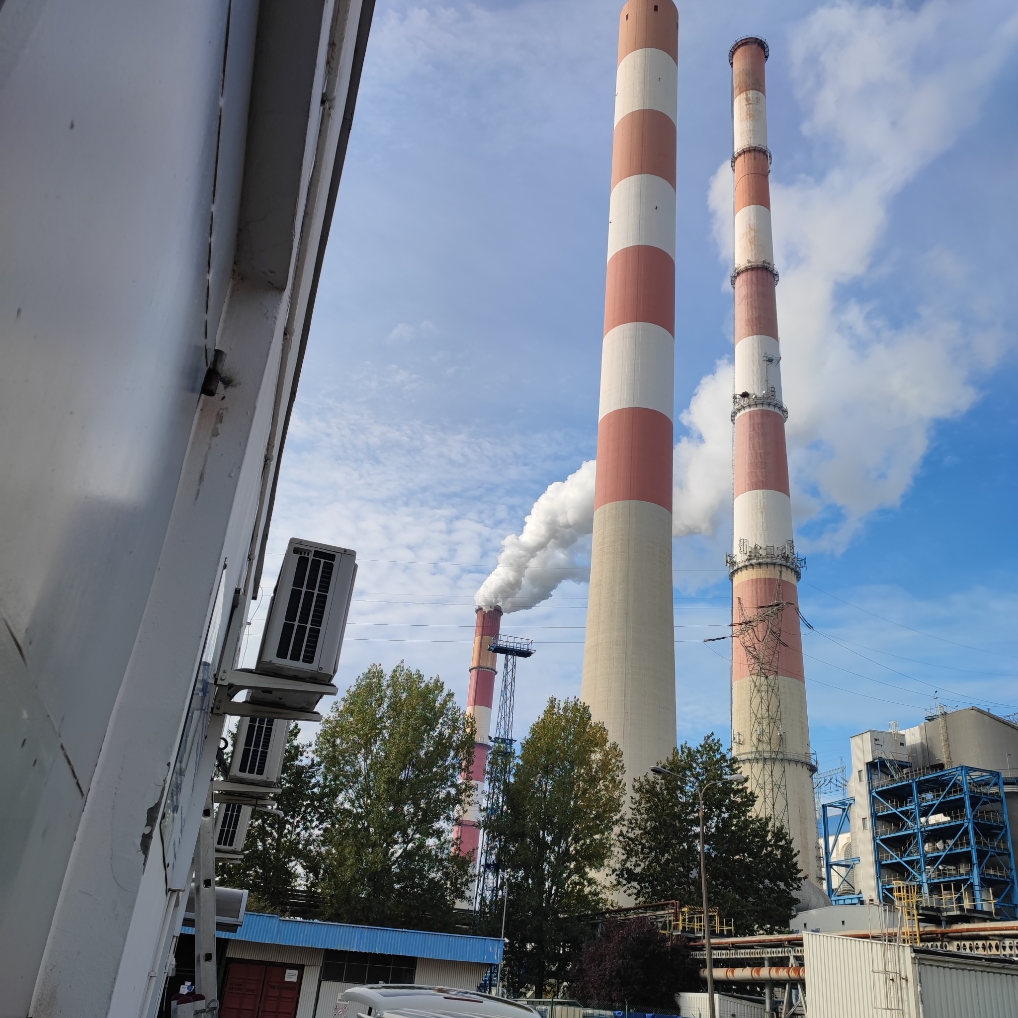 Dwa wysokie kominy elektrowni z czerwono-białymi pasami, z jednego wydobywa się biały dym, na pierwszym planie fragment budynku z zamontowanymi klimatyzatorami.
