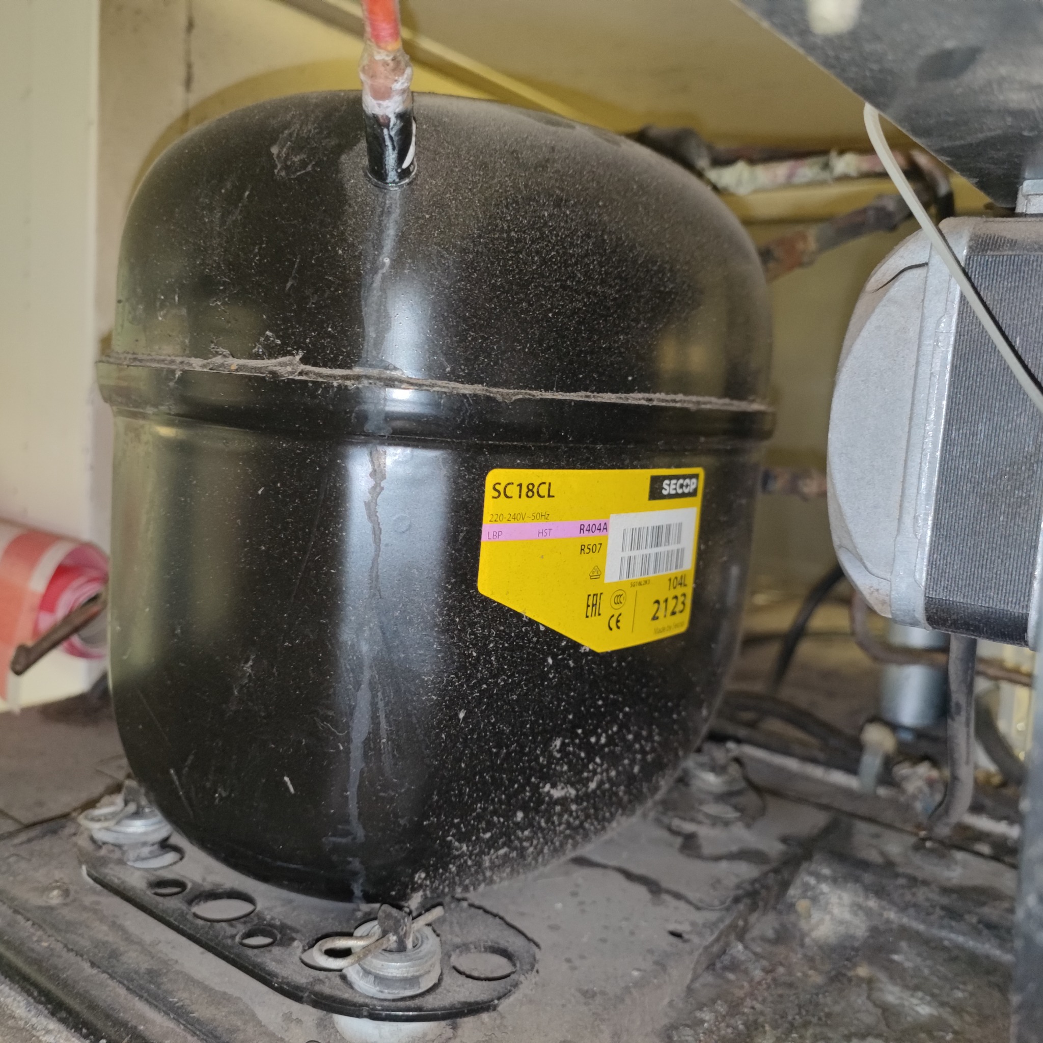 Czarny, cylindryczny kompresor SC18CL firmy Secop, z żółtą naklejką informacyjną, zamontowany na metalowej podstawie, widoczne ślady kurzu i zabrudzeń.