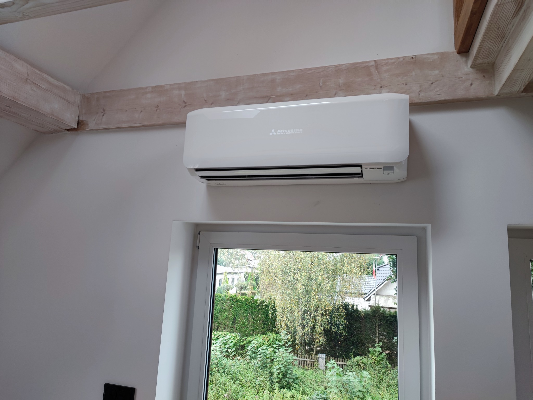 Biały klimatyzator Mitsubishi zamontowany na ścianie pod drewnianą belką stropową, widok z okna na zieleń.