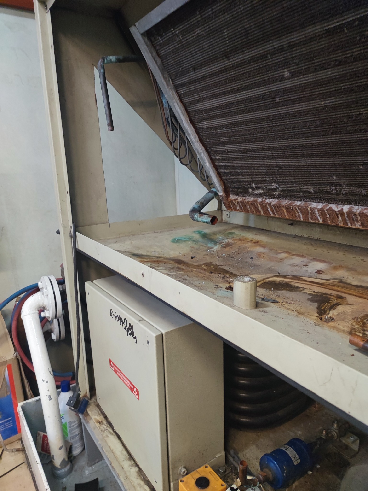 Zdemontowany, brudny wymiennik ciepła z miedzianymi rurkami, widoczne ślady korozji i zabrudzenia, obok skrzynka z napisem R-404A-2,8Kg i ostrzeżeniem.