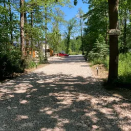 Długa, żwirowa droga dojazdowa, oświetlona niskimi lampami, prowadząca do domu widocznego w oddali, z czerwonym samochodem zaparkowanym obok, otoczona drzewami i tabliczką informującą o terenie...