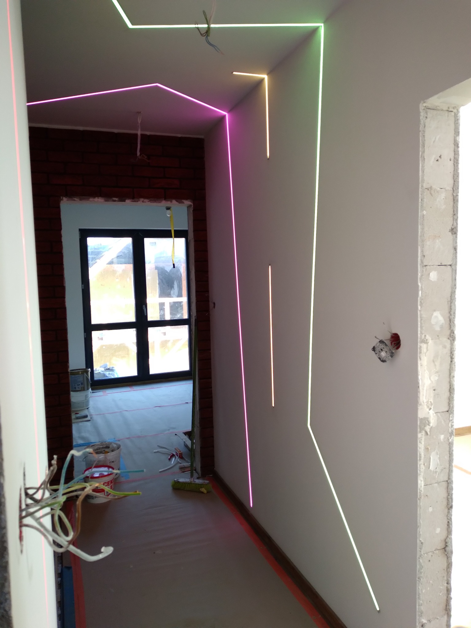 Wnętrze w trakcie wykańczania z widocznymi liniami lasera wyznaczającymi poziom na ścianach i suficie, surowy ceglany mur w tle, widoczne kable elektryczne i narzędzia malarskie.
