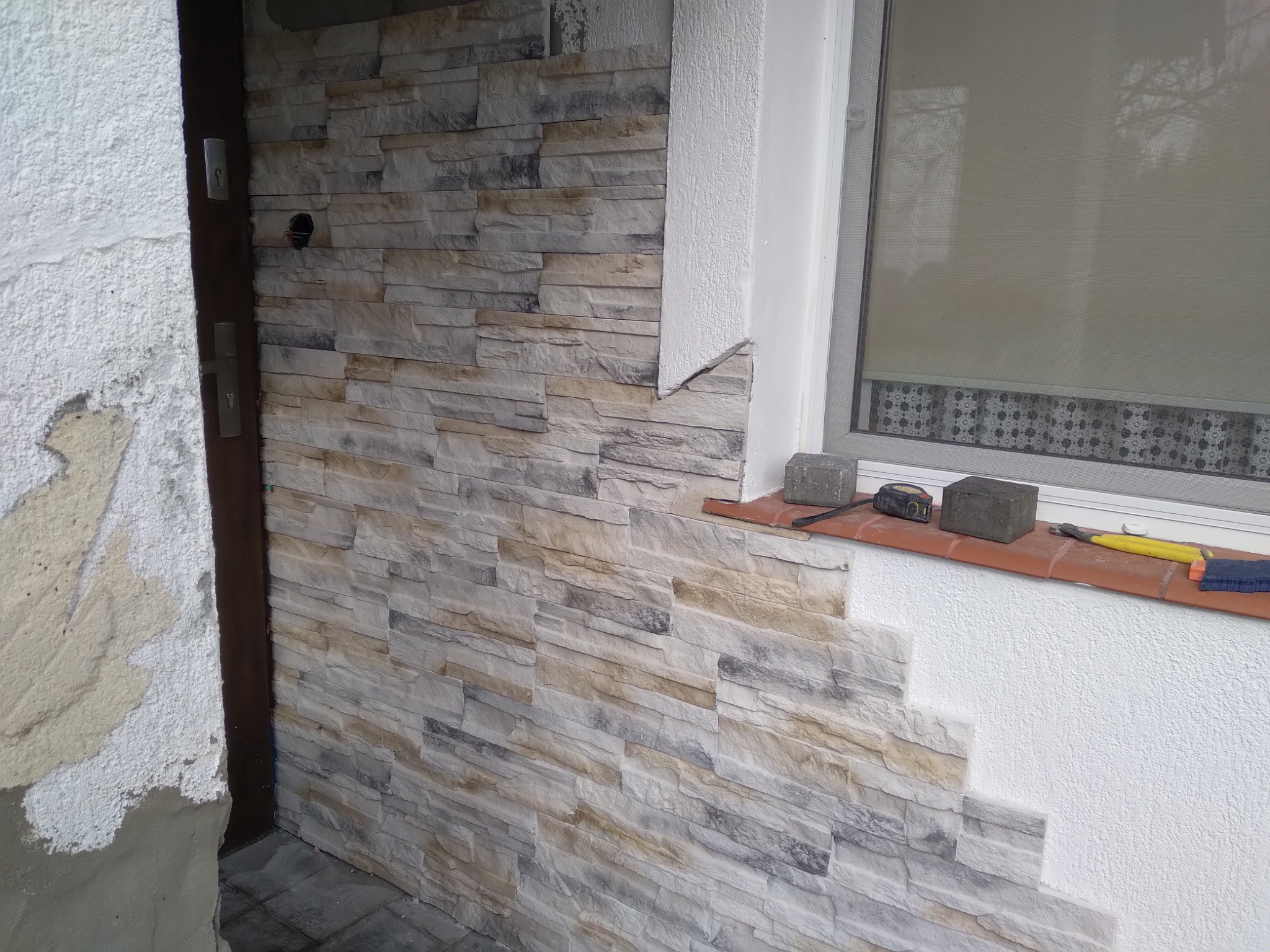 Montaż dekoracyjnej okładziny kamiennej na elewacji budynku, widoczne narzędzia pracy i fragment okna.