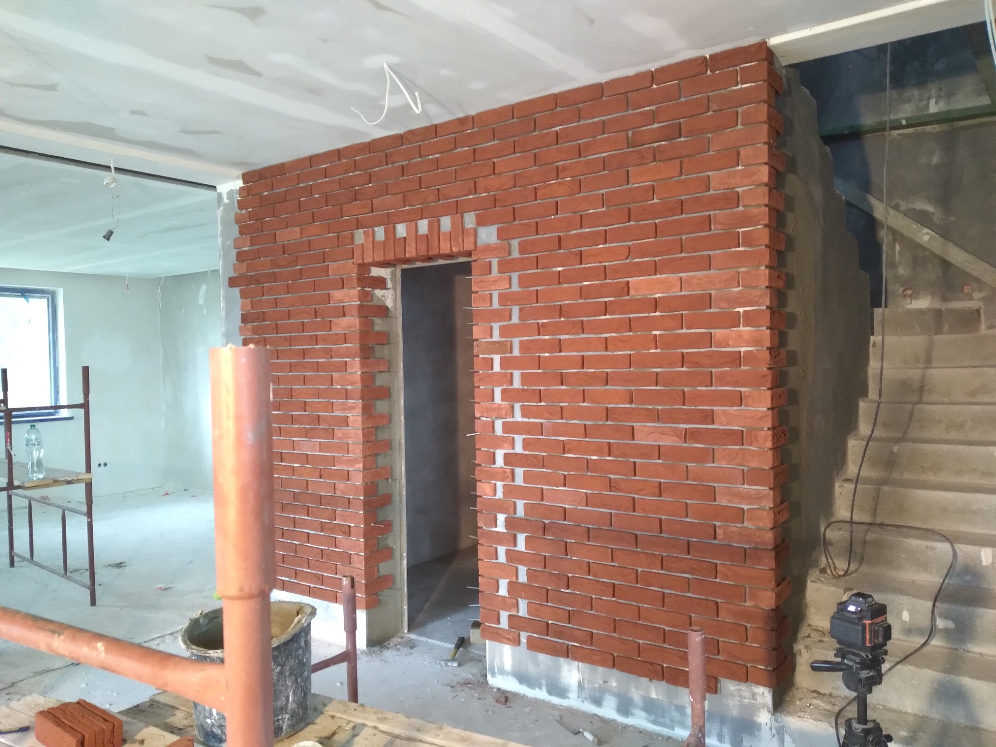 Murowana ściana z czerwonej cegły klinkierowej w trakcie budowy, widoczne spoiny, fragment ościeżnicy drzwiowej, narzędzia budowlane i laser krzyżowy na statywie w surowym wnętrzu.