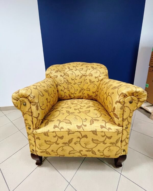 Żółty fotel klubowy z brązowym roślinnym wzorem, ozdobne nity na podłokietnikach, na tle ściany w kolorach białym i granatowym.