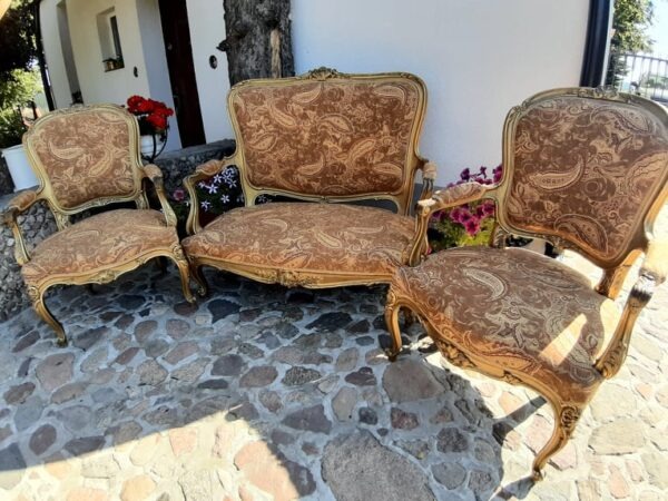 Zestaw mebli w stylu Ludwika: sofa dwuosobowa i dwa fotele z tapicerką w brązowe wzory paisley, ustawione na kamiennej posadzce przed budynkiem.