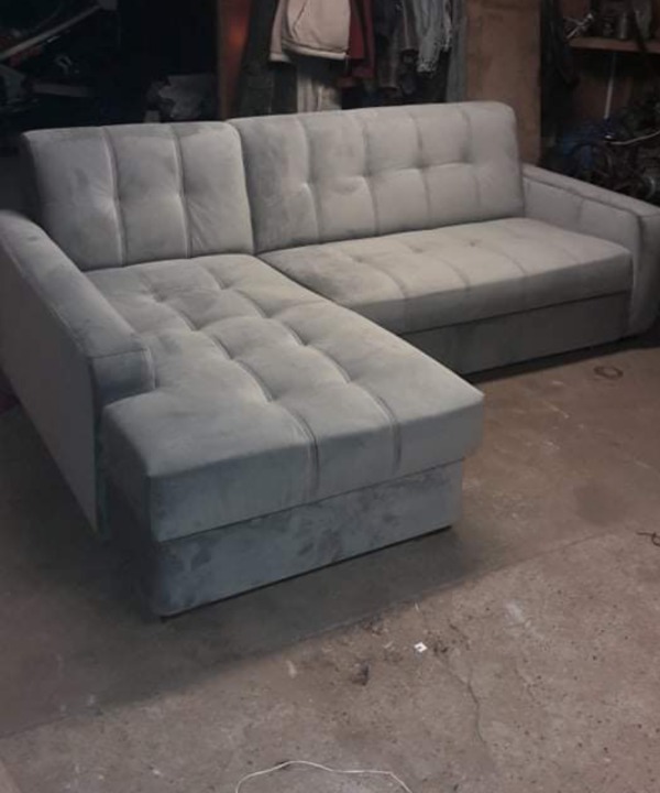 Szara sofa narożna z pikowanymi siedziskami i oparciami, sfotografowana w warsztacie z betonową podłogą i widocznymi narzędziami w tle.