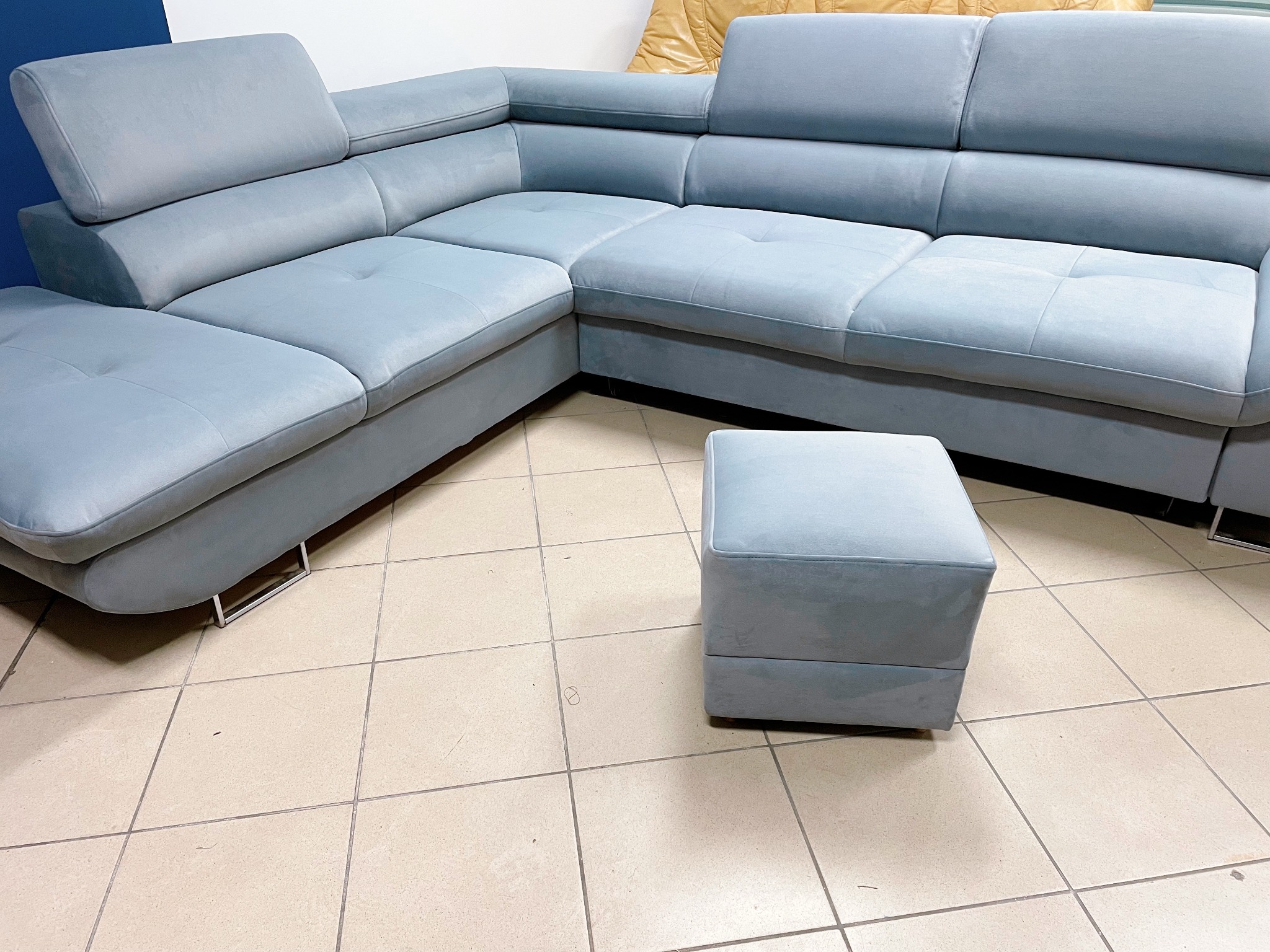 Narożnik tapicerowany w odcieniach błękitu z pufą na kafelkowej podłodze, widoczne metalowe nóżki
