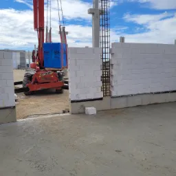 Czerwony podnośnik teleskopowy transportuje pakiet białych bloczków Silka na placu budowy, gdzie widać fragment ściany z tych bloczków oraz zbrojenie pionowe, a w tle niebo z chmurami.