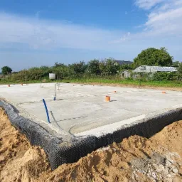 Wylana płyta fundamentowa z hydroizolacją pionową, widoczne wystające rury instalacyjne, teren budowy z piaskiem i w oddali zabudowania gospodarcze.