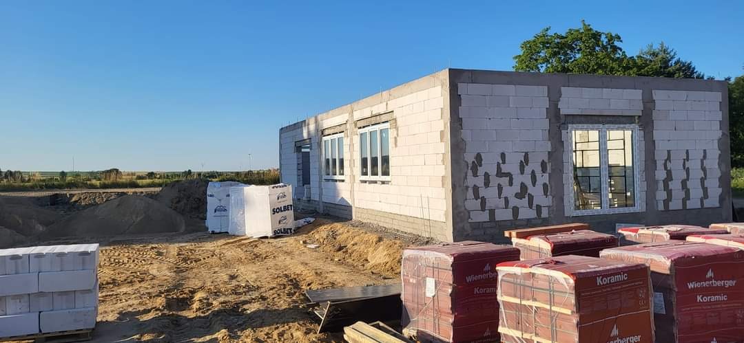 Niedokończony budynek z białych bloczków, z widocznymi oknami i stertami cegieł Koramic oraz bloczków Solbet na placu budowy.