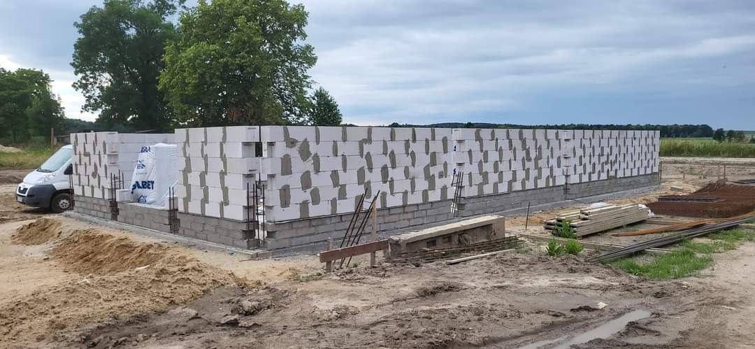 Wznoszenie ścian parteru domu z betonu komórkowego na fundamencie z bloczków betonowych; widoczny częściowo zaparkowany biały samochód dostawczy i materiały budowlane.