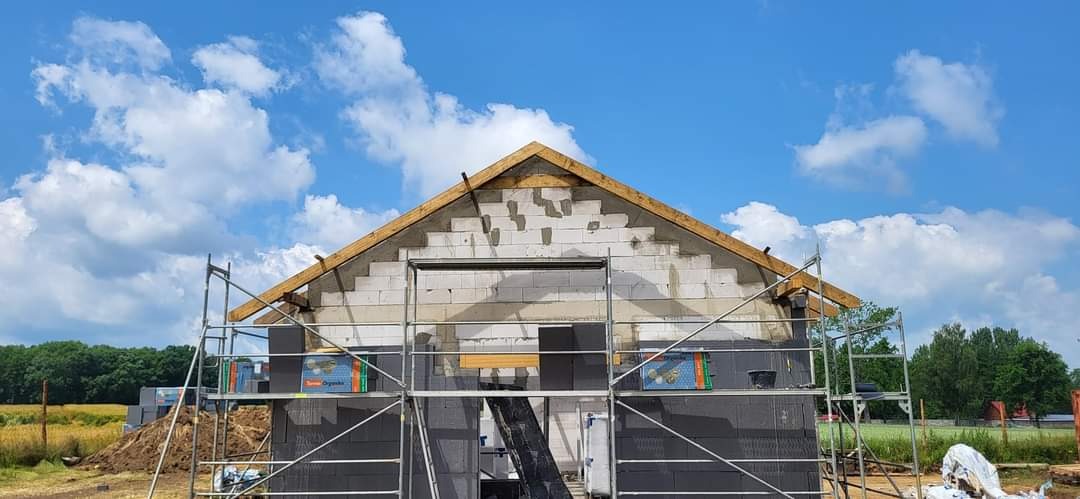 Wznoszona ściana szczytowa budynku z białych bloczków, częściowo ocieplona czarnym styropianem, widoczne rusztowanie i drewniana konstrukcja dachu na tle błękitnego nieba z chmurami.