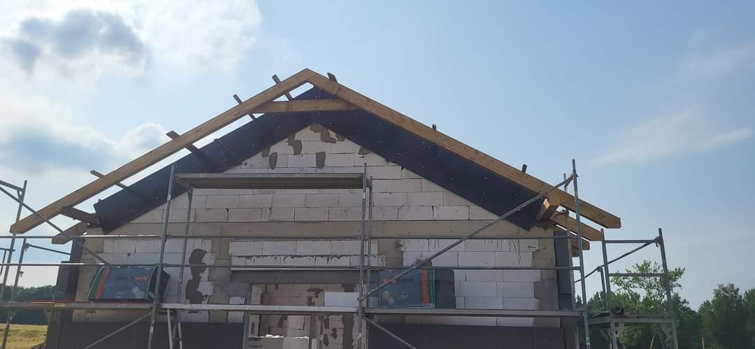 Wznoszona ściana domu z betonu komórkowego z widocznym drewnianym szkieletem dachu i fragmentami czarnej membrany, otoczona rusztowaniem.