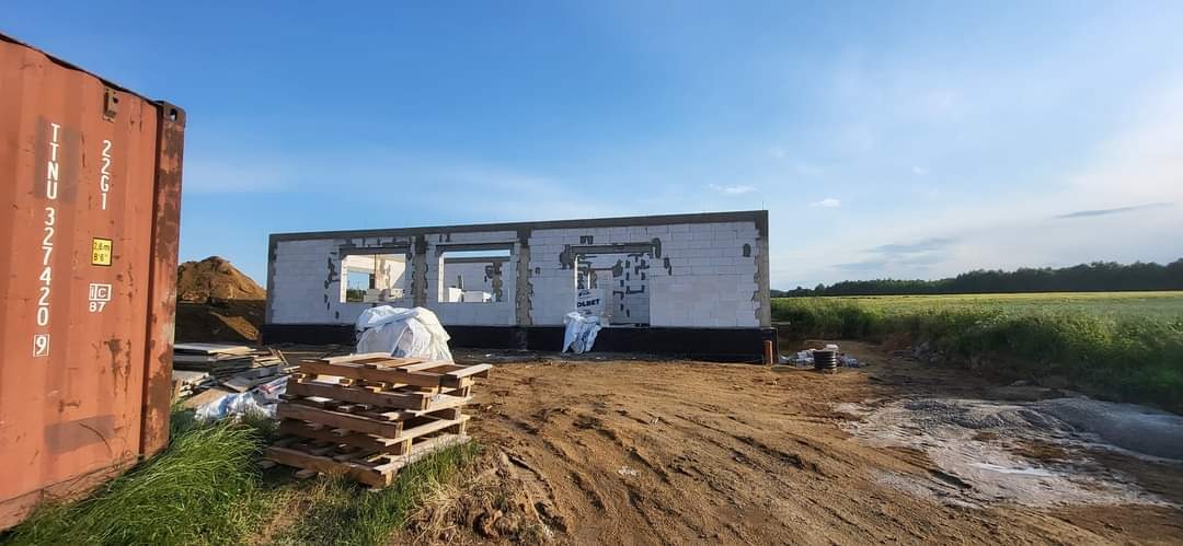 Budowa domu jednorodzinnego w trakcie wznoszenia ścian z bloczków betonu komórkowego, widoczny kontener budowlany, paleta z materiałami budowlanymi i przygotowany teren pod budowę.
