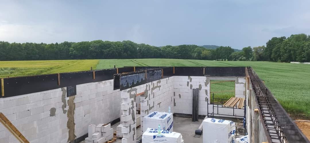 Budowa domu jednorodzinnego w stanie surowym otwartym: widok na ściany z betonu komórkowego z czarnym szalunkiem, zbrojenie wieńca na tle zielonego pola i lasu.