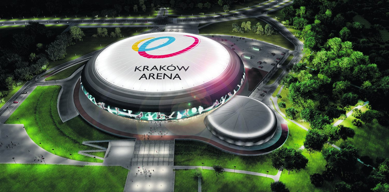 Kraków Arena z lotu ptaka, widoczne wejście główne, parking, zieleń parkowa i oświetlenie nocne, na dachu logo obiektu.