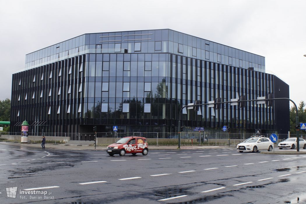 Nowoczesny, przeszklony budynek biurowy z czarną elewacją i częściowo otwartymi oknami, widziany z perspektywy ulicy w pochmurny dzień. Na pierwszym planie mokra jezdnia z przejeżdżającymi...