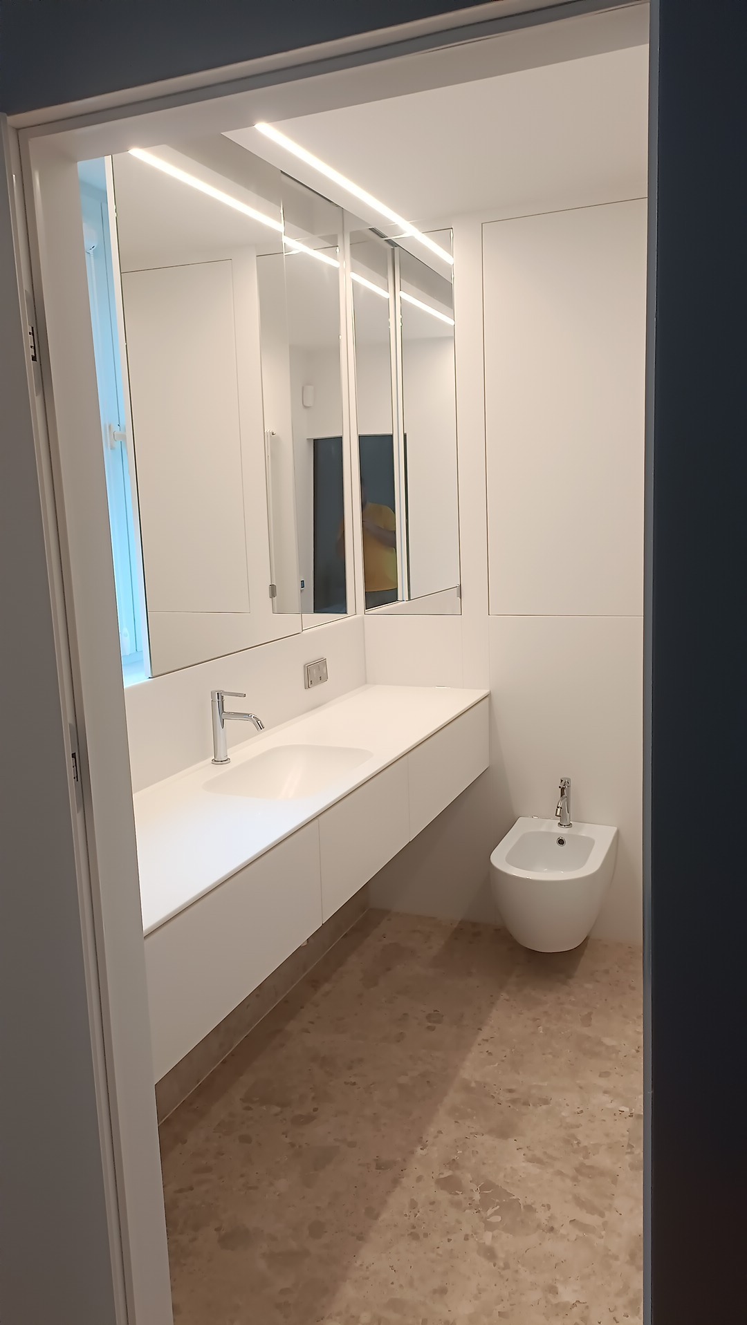 Nowoczesna łazienka z białymi szafkami pod umywalką, trzema lustrami z oświetleniem LED, białym bidetem i beżową marmurową podłogą widziana zza framugi drzwi.