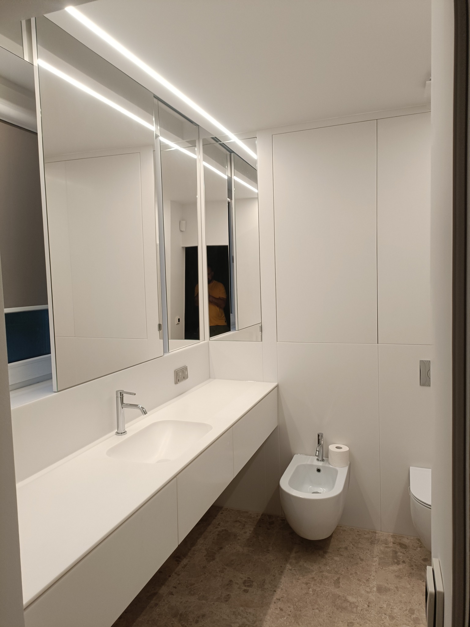 Nowoczesna łazienka w bieli z podświetlanym lustrem, umywalką nablatową, bidetem i toaletą. Na podłodze marmurowe płytki w odcieniach brązu.