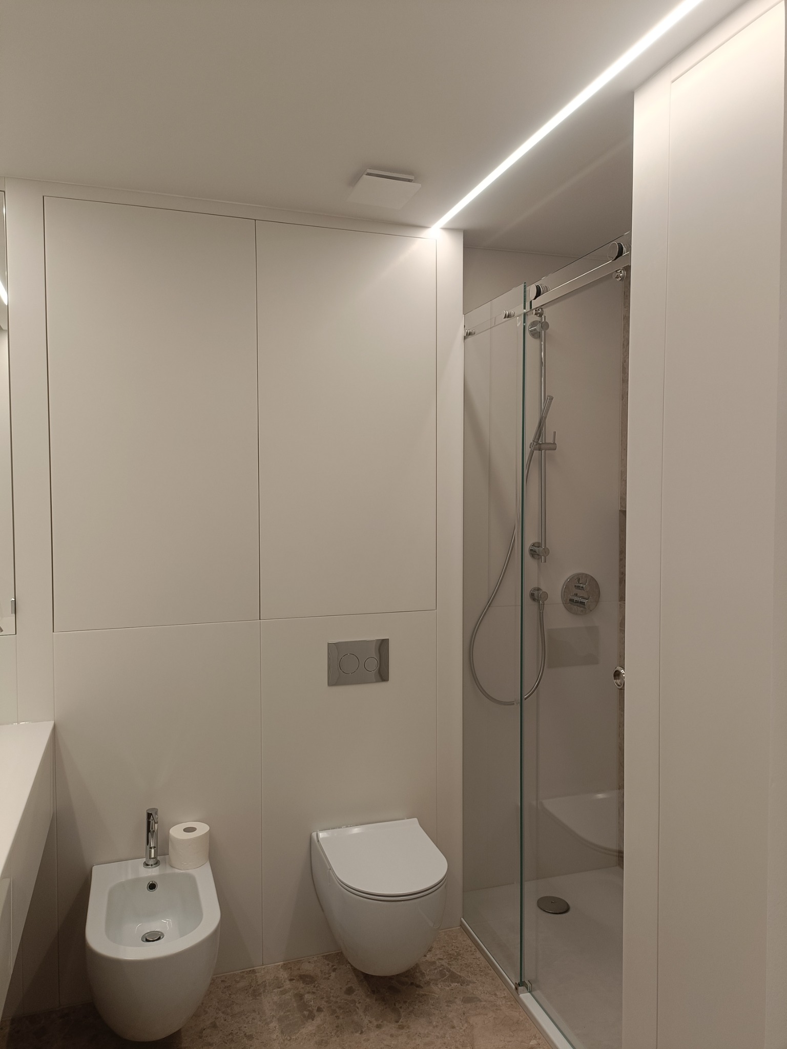 Nowoczesna łazienka z białymi szafkami, podwieszaną toaletą, bidetem z rolką papieru, kabiną prysznicową ze szklanymi drzwiami i oświetleniem LED podsufitowym.