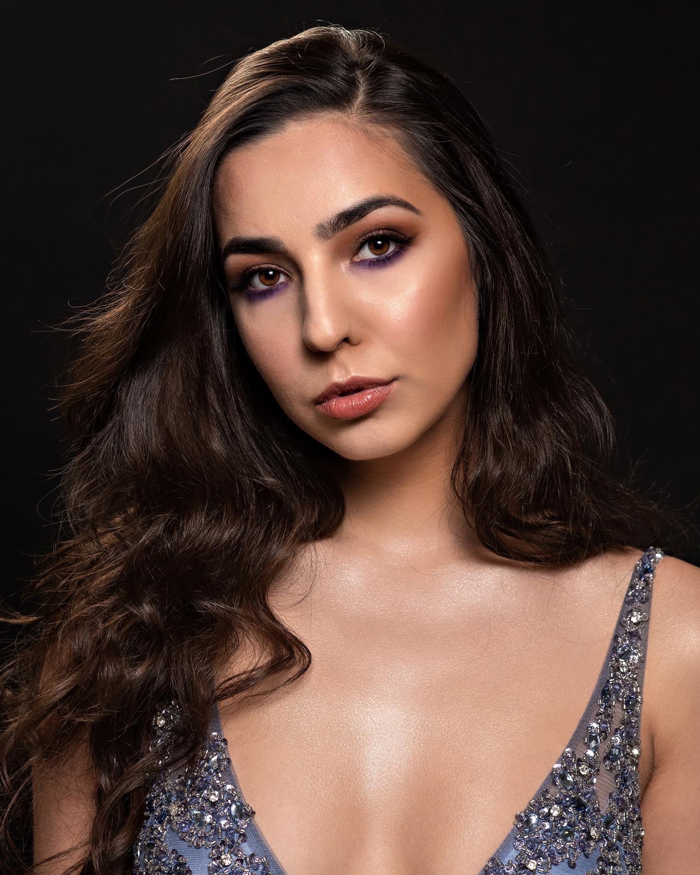 Portret kobiety z profesjonalnym makijażem oczu w odcieniach fioletu i brązu, delikatnym połyskiem na ustach i luźno opadającymi, falowanymi, ciemnymi włosami na czarnym tle, ubrana w sukienkę...