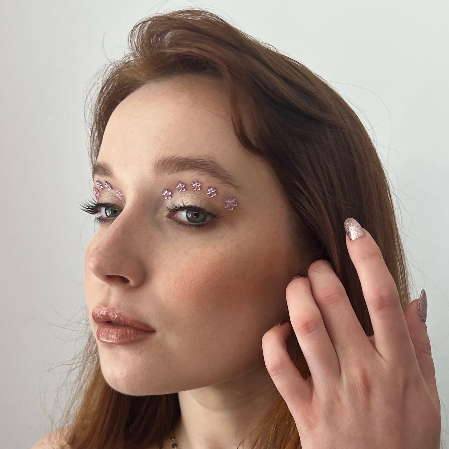 Portret kobiety z subtelnym, artystycznym zdobieniem w postaci różowych kwiatków nad brwiami, błyszczącymi ustami i brokatowymi paznokciami, skierowana w lewo.