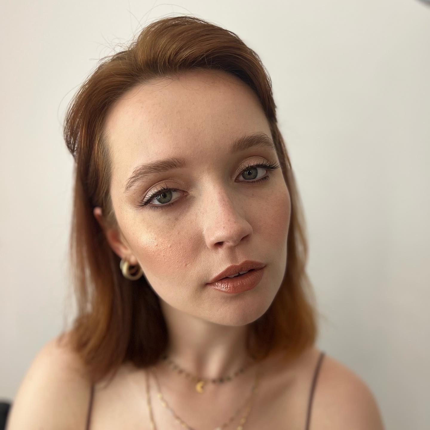 Portret kobiety z delikatnym makijażem: rozświetlona cera, brązowe cienie na powiekach, błyszczące usta w odcieniu nude, złote kolczyki i naszyjniki.