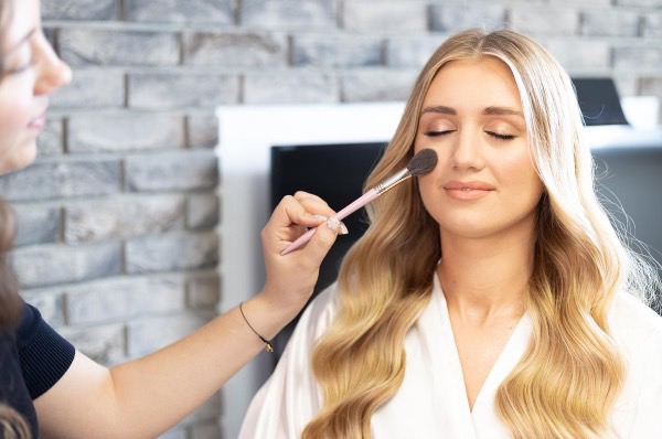 Aplikacja pudru pędzlem na policzek blondwłosej kobiety w białym szlafroku, w tle ściana z cegły. Delikatny, dzienny makijaż.