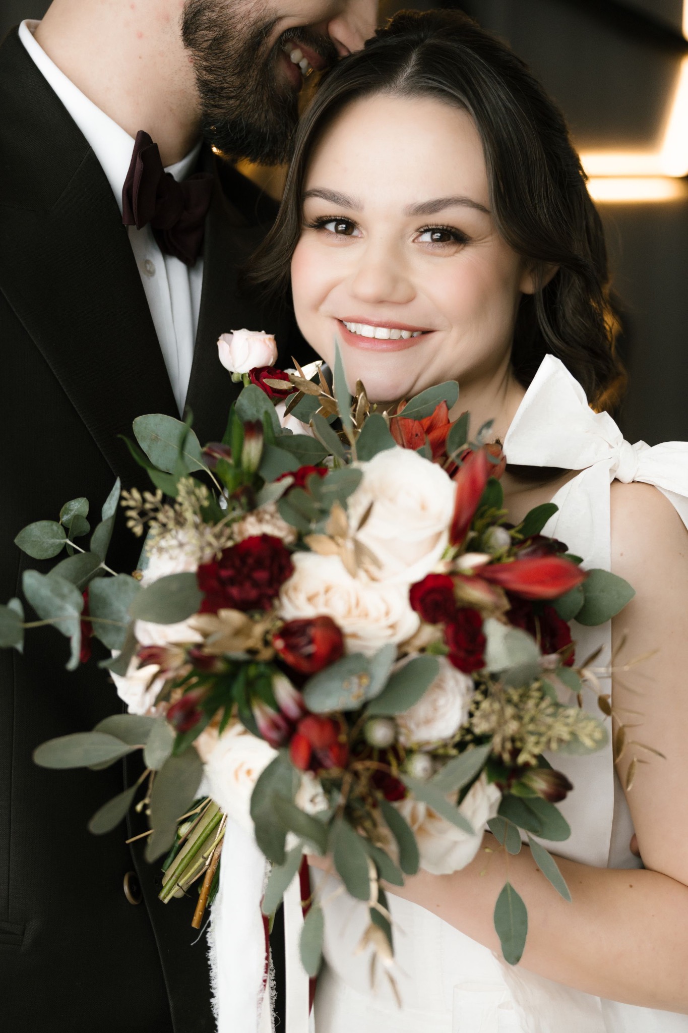 Uśmiechnięta panna młoda z bukietem ślubnym, widoczny fragment garnituru pana młodego z bordową muchą. Makijaż ślubny podkreślony, delikatny.