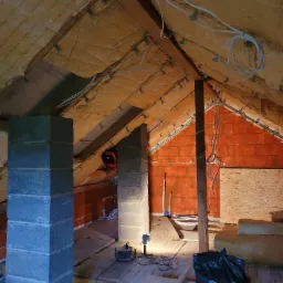 Poddasze w trakcie prac: ocieplenie wełną mineralną poddasza, widoczne krokwie, instalacja elektryczna, komin z bloczków betonowych, fragment ściany z czerwonej cegły oraz narzędzia i materiały...