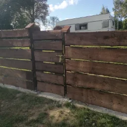 Nowoczesne, poziome ogrodzenie z ciemnego drewna z zamykaną furtką na tle przyczepy kempingowej i drzew.