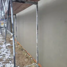 Świeżo otynkowana ściana budynku w trakcie prac wykończeniowych, widoczne rusztowanie i resztki śniegu na gruncie.