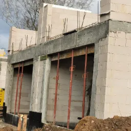 Wznoszenie ścian parteru domu z betonu komórkowego, widoczne stalowe pręty zbrojeniowe i podpory szalunkowe, ziemia w wykopie.