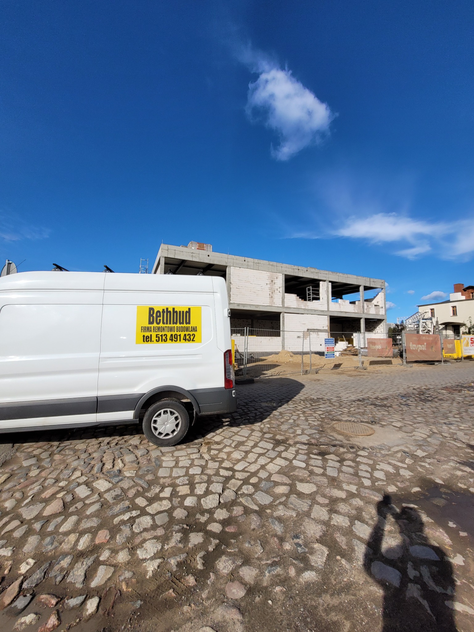 Budynek w trakcie wznoszenia z białych bloczków, widoczny zza zaparkowanego białego busa z żółtym logo firmy budowlanej na tle brukowanej ulicy i błękitnego nieba z pojedynczą chmurą.