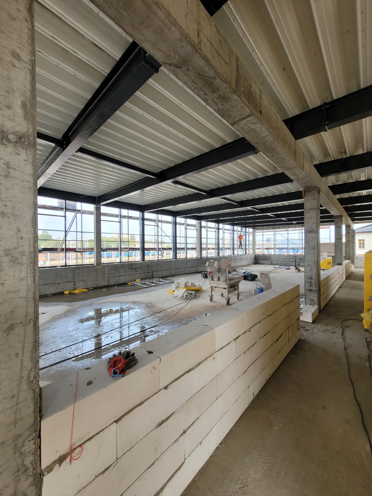 Wnętrze budynku w trakcie budowy, widoczne ściany z bloczków, stalowa konstrukcja dachu, betonowe słupy, okna i narzędzia budowlane.