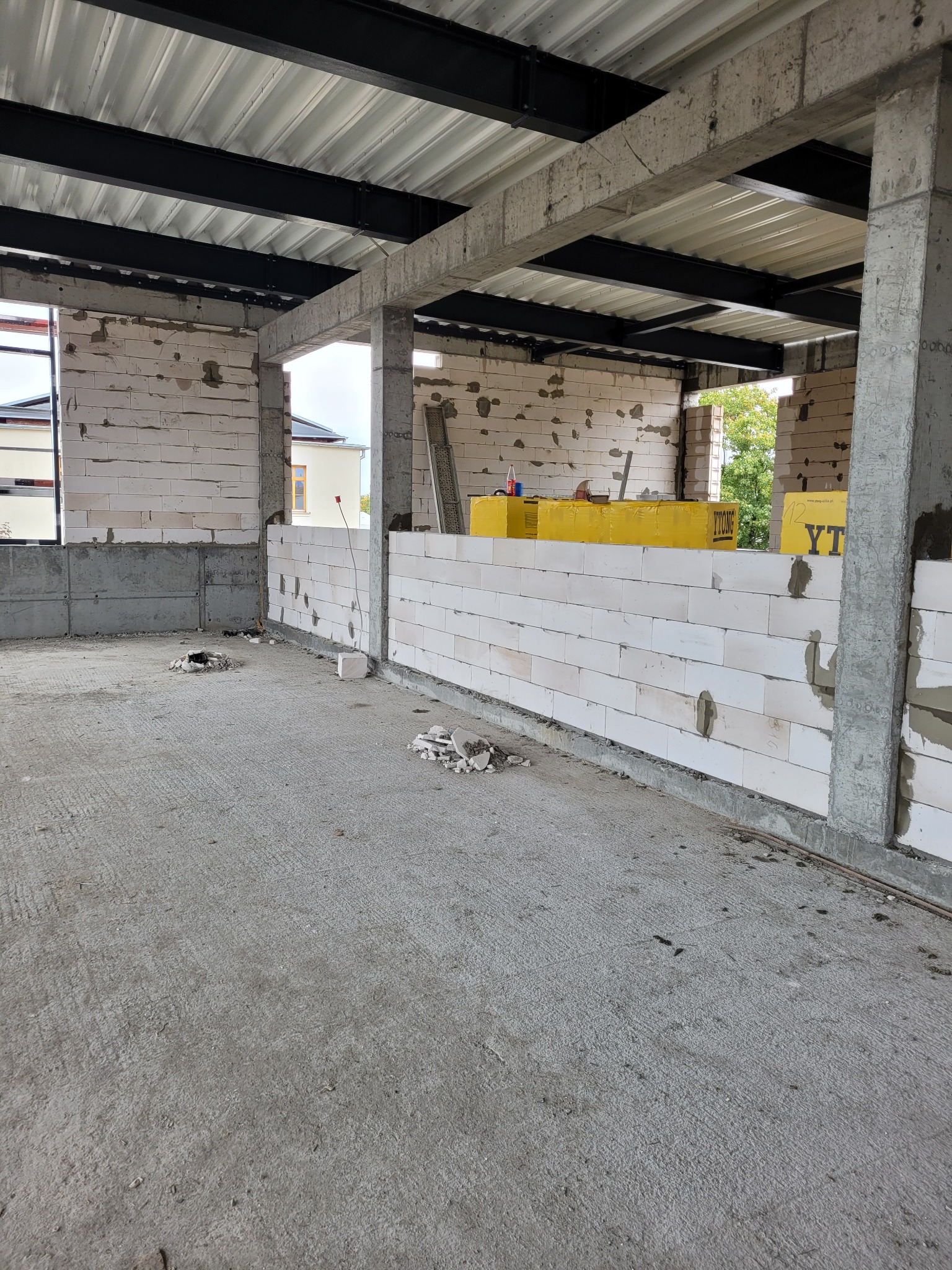 Surowy stan domu w trakcie budowy, ściany z białych bloczków betonowych, betonowe słupy konstrukcyjne, stalowe belki stropowe, widoczne otwory w posadzce i gruz.
