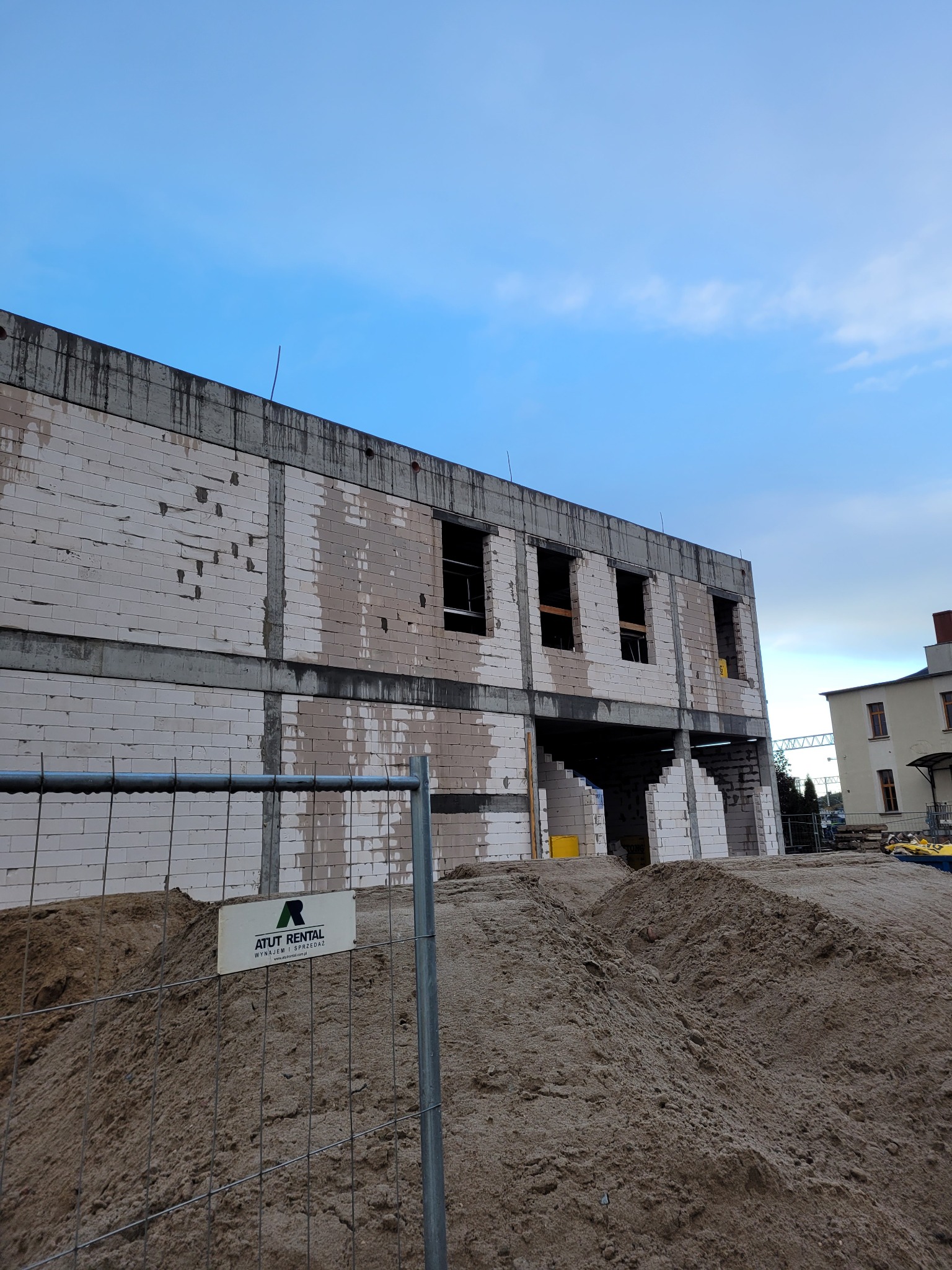 Wznoszona dwukondygnacyjna konstrukcja budynku z białych bloczków, widoczne otwory okienne bez ram, betonowe nadproża i słupy, na pierwszym planie pryzma piasku i tymczasowe ogrodzenie z tablicą...