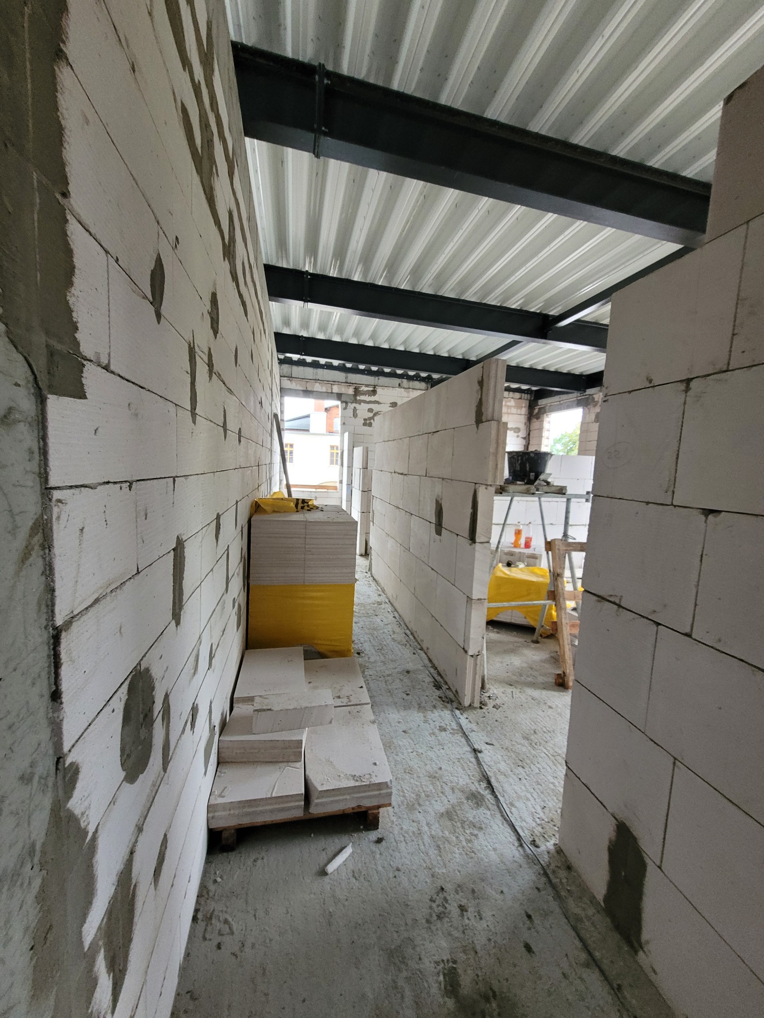 Wnętrze budynku w trakcie budowy, ściany z białych bloczków, widoczne zapasy materiałów budowlanych i rusztowanie z wiadrem.