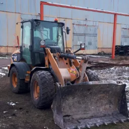 Pomarańczowo-czarna ładowarka kołowa CAT z brudną łyżką na tle szarego budynku przemysłowego z czerwoną konstrukcją rurową.