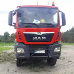 Czerwona ciężarówka MAN z żółtym kaskiem i dokumentami za szybą, stojąca na piaszczystym terenie.