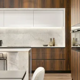 Nowoczesna kuchnia z drewnianymi szafkami, marmurowym blatem i wyspą z hokerami, oświetlona listwą LED pod szafkami wiszącymi.