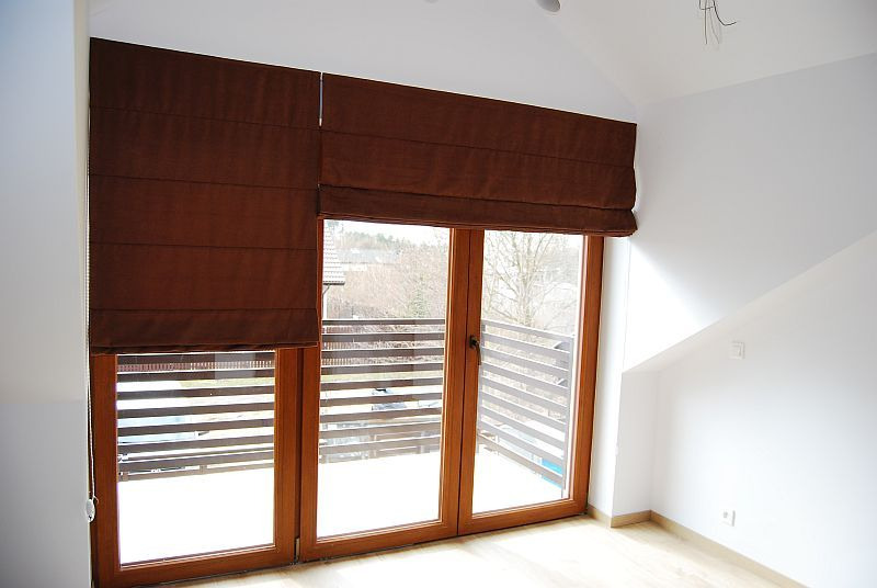 Brązowe tekstylne osłony okienne zamontowane w oknach balkonowych z drewnianymi ramami, widok na balkon z drewnianą balustradą i drzewa w tle.