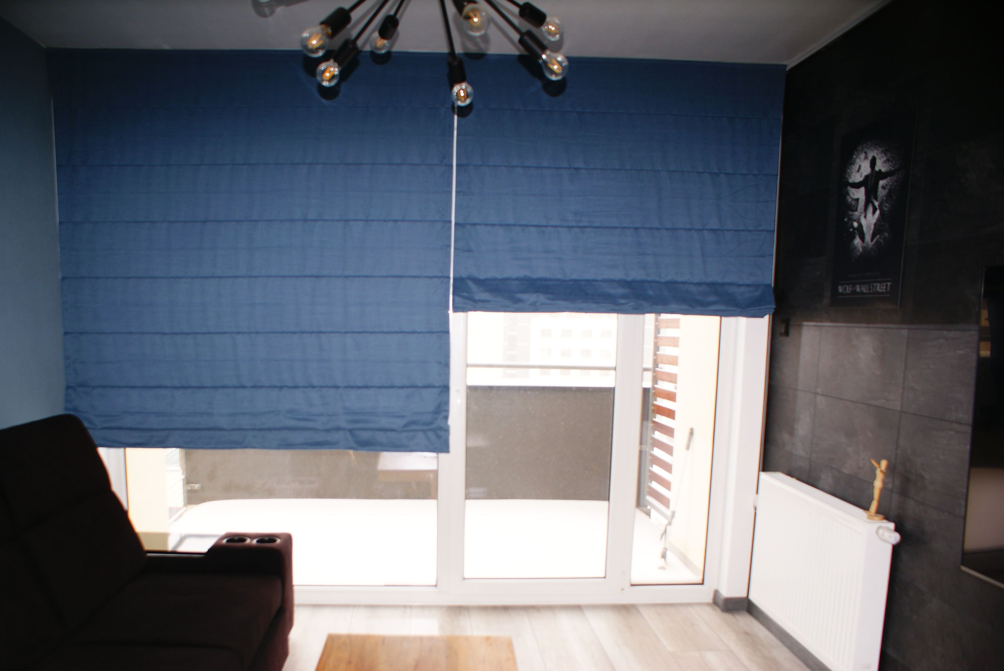 Niebieskie tekstylne osłony okienne w nowoczesnym salonie z wyjściem na balkon, jedna opuszczona częściowo, widoczny fragment sofy i lampa sufitowa.