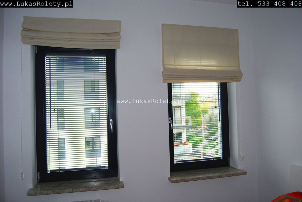Dwa okna z zamontowanymi roletami rzymskimi w kolorze kremowym, jedno z żaluzjami aluminiowymi za roletą, widok na budynek i zieleń za oknami.