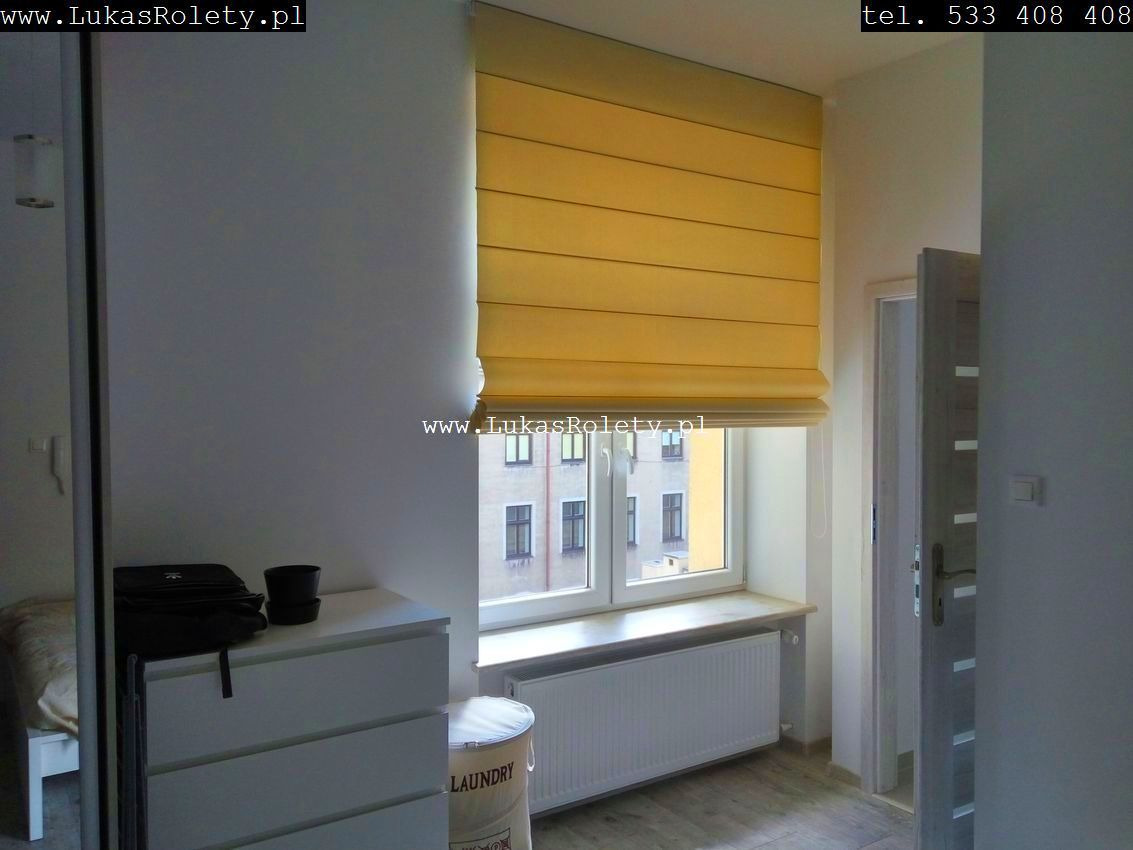 Żółta, poziomo plisowana osłona okienna zamontowana w oknie z widokiem na budynek mieszkalny, widoczny fragment pokoju z grzejnikiem i komodą.